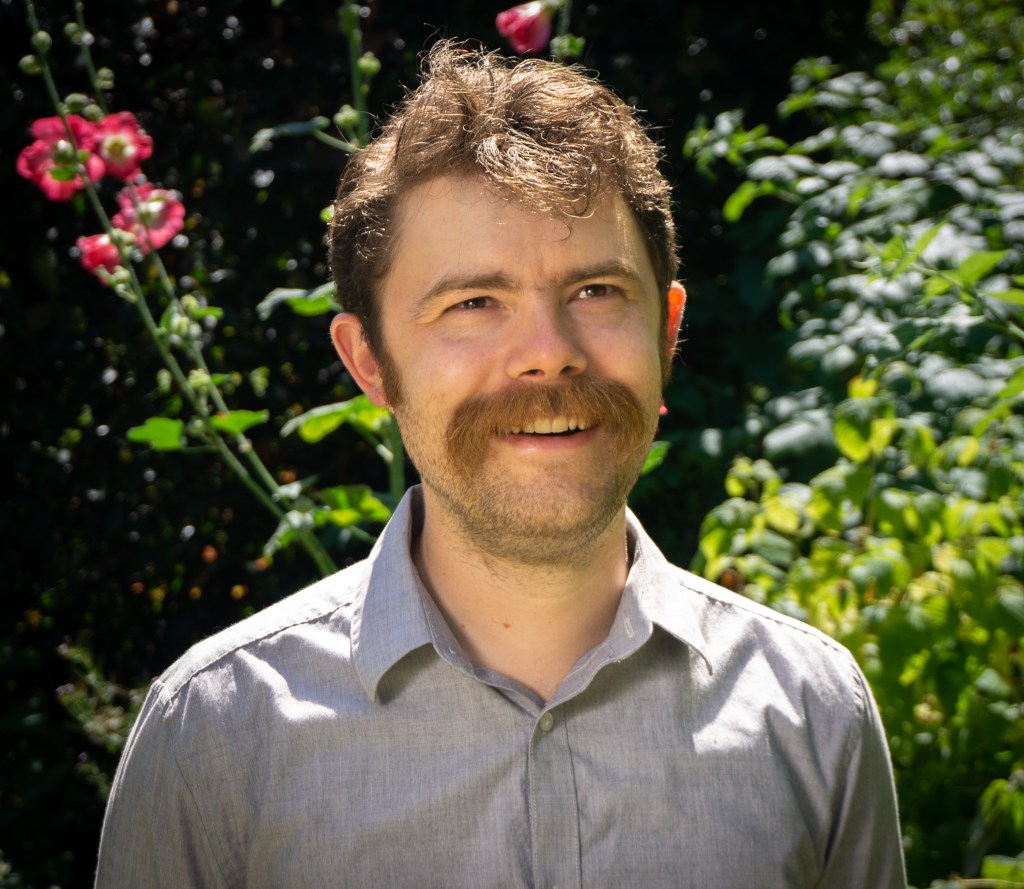 Headshot of a man with a mustache in front of greenery and flowers.
